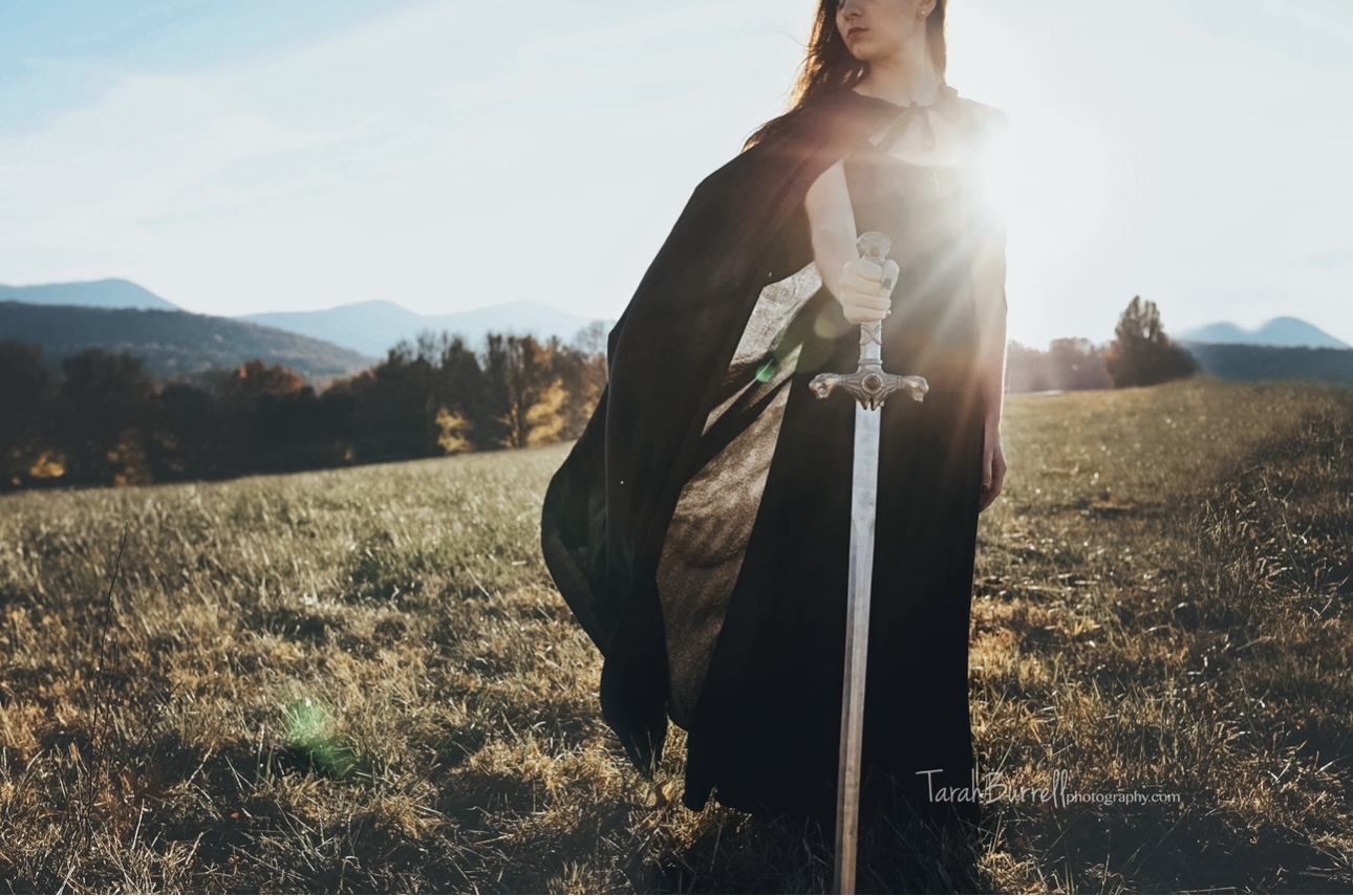 A woman stands in a field, wearing a black dress or tunic and holding large pointed silver sword in front of her. The sun ...