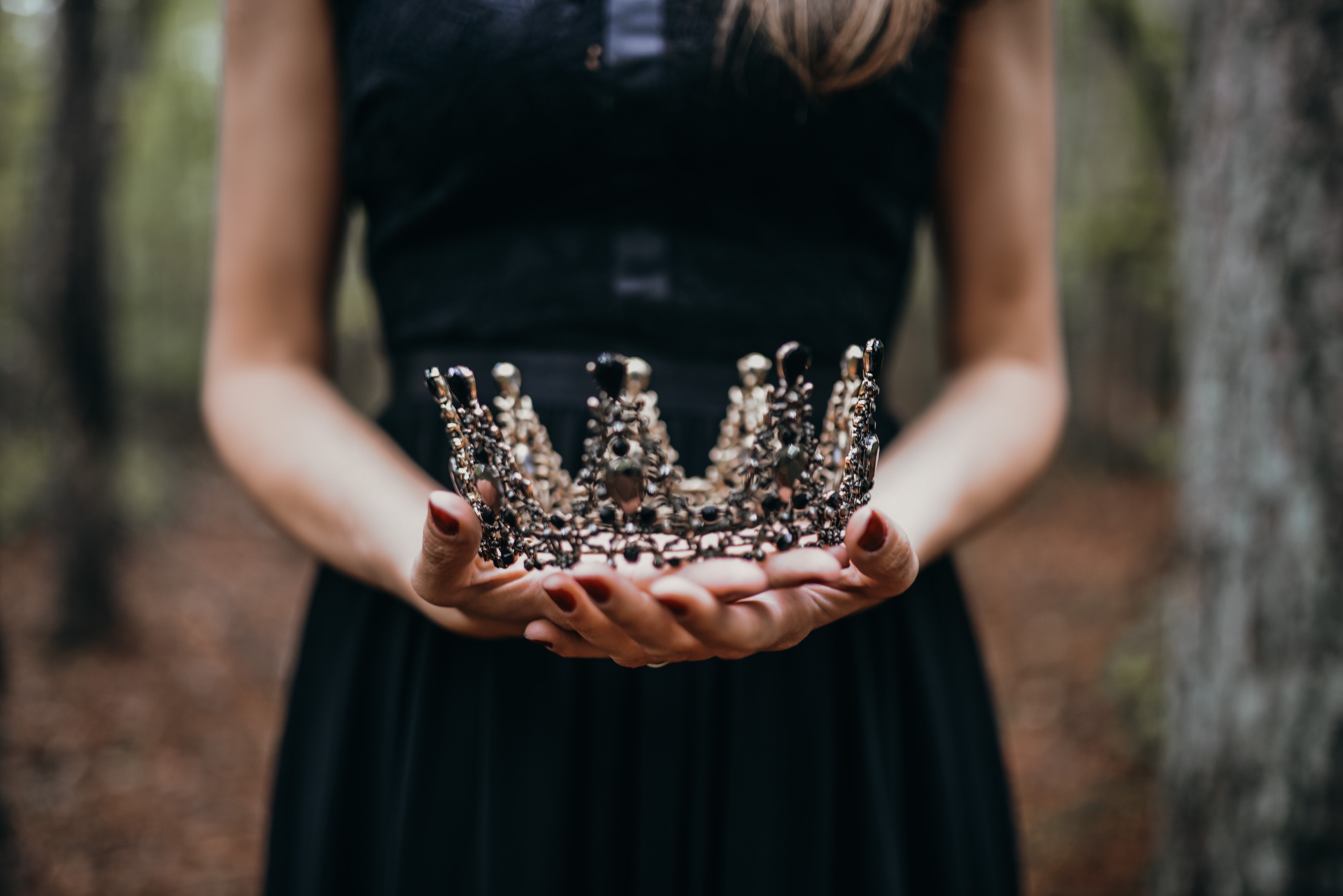 A woman in a black dress holds a gold crown with gems, standing in front of a leaf-strewn forest floor, with out-of-focus ...